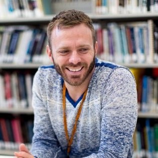 A guy with a blue sweater smiling with books in the background