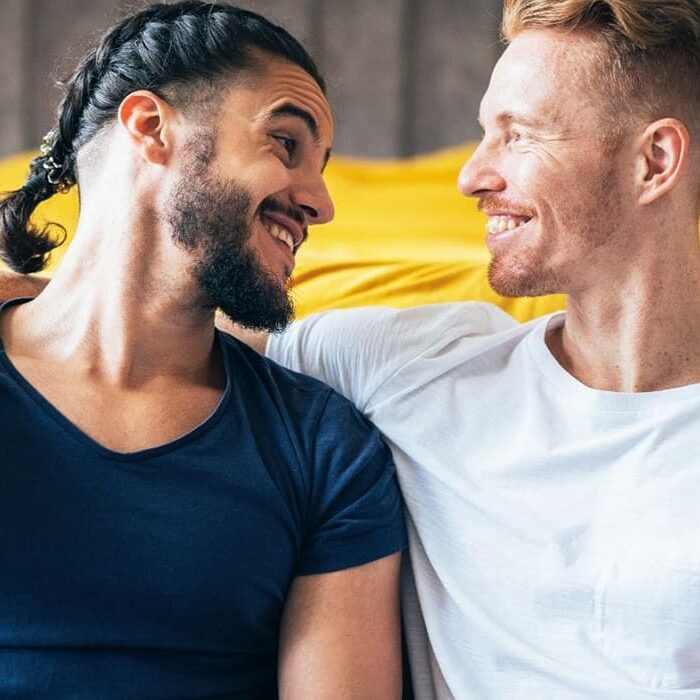Two guys, one with a braid in their hair smiling at each other on a yellow couch