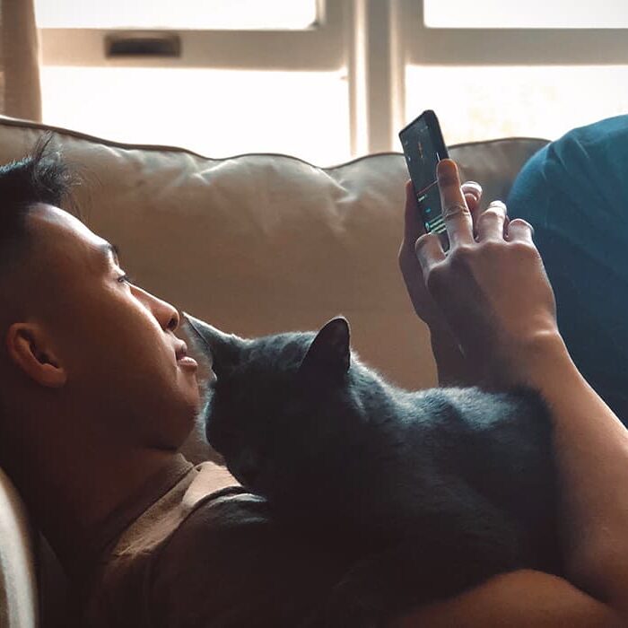 A young guy lying down on a couch, holding a cat while on their phone