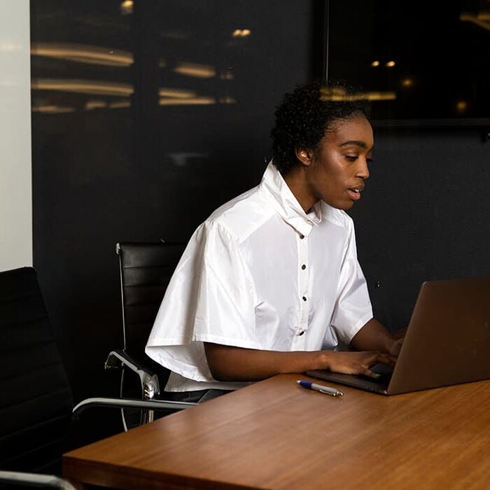 A black person wearing a white button up shirt sitting in a board room working on a gray laptop