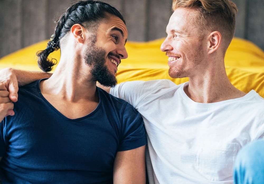 Two guys, one with a braid in their hair smiling at each other on a yellow couch