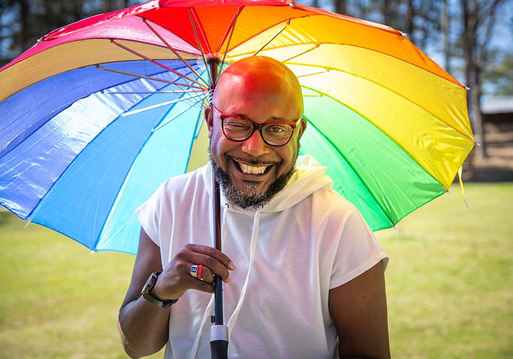 A person standing under a rainbow umbrella and winking at the camera
