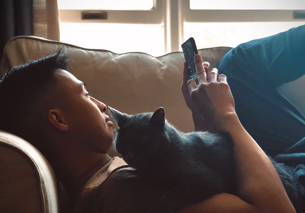 A young guy lying down on a couch, holding a cat while on their phone