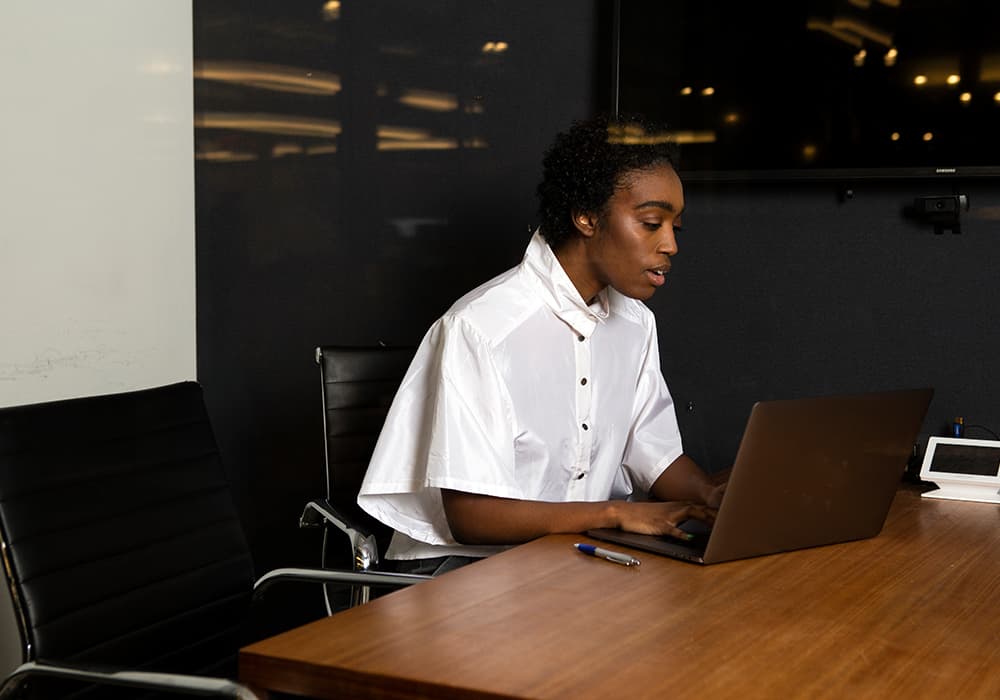 A black person wearing a white button up shirt sitting in a board room working on a gray laptop