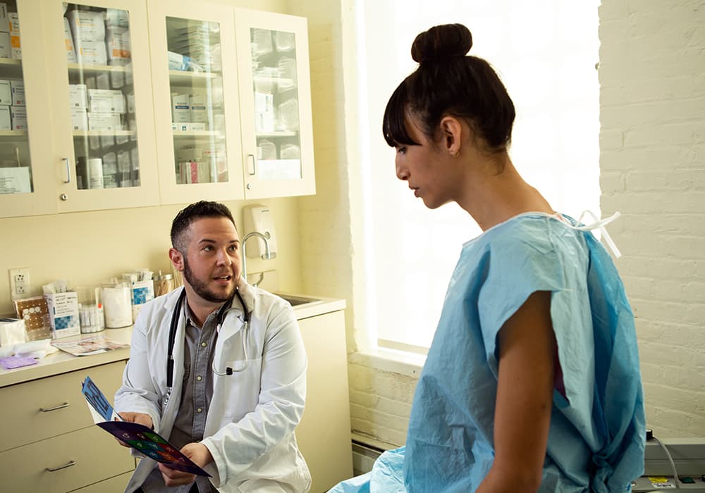 A doctor in a medical room sharing results with patient who has black hair in a bun with a blue gown on